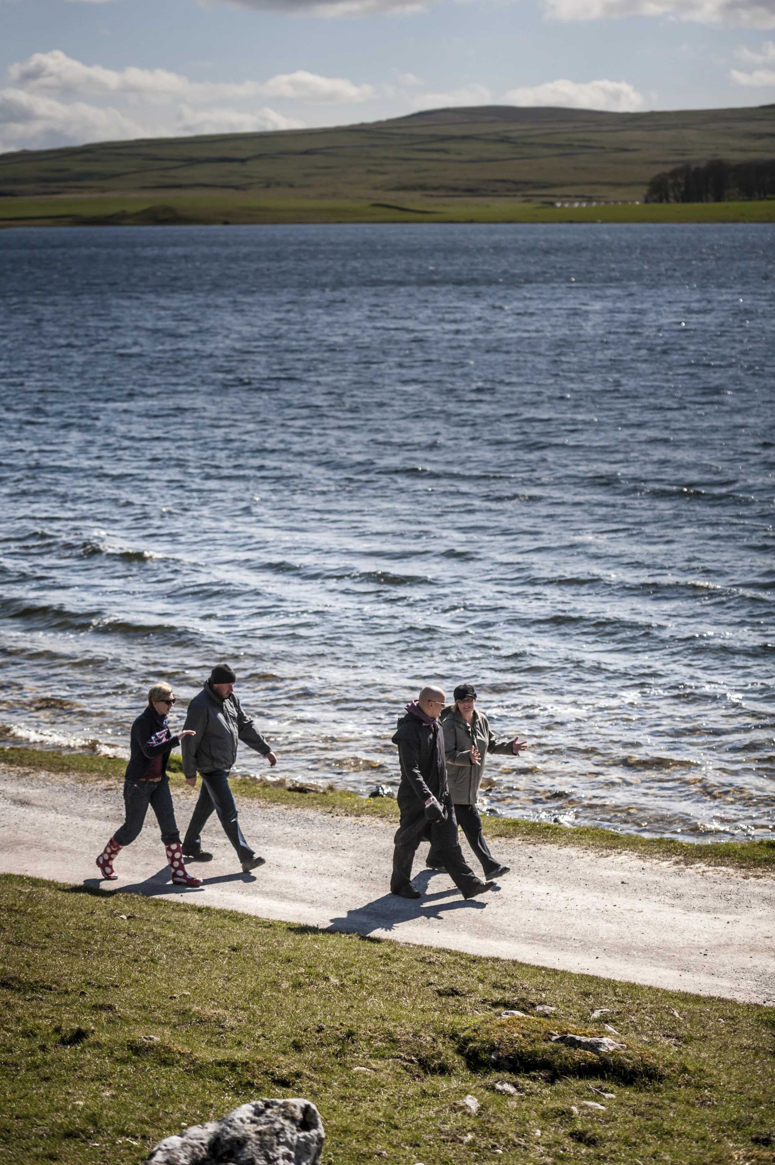 Four people walking along a path beside a large lake, with hills and cloudy sky in the background; cool blue and green tones